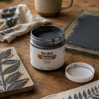 Encre de bloc Tri-Art gris foncé en pot ouvert avec tampon feuille gravée et échantillons de tissu